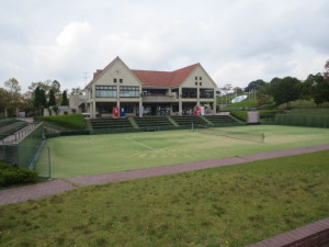 前方に2つの三角屋根をしたクラブハウスがあり、手前に人工芝のテニスコートがある、しあわせの村センターコート施設をコート側から写した写真