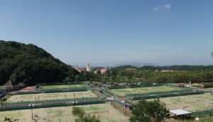 後方に小高い山があり、フェンスで囲まれたテニスコートが隣接している、しあわせの村テニスコートを高台から写した写真