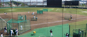 マウンドからボールを投げる選手、バットを持って構えている選手、内野や外野を守っている選手と、キャッチャーの後ろに立っている審判がいる練習中のG7スタジアム神戸の写真